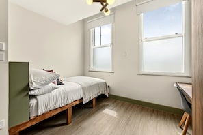 Ein kleines Zimmer mit einem Einzelbett, Holzboden, zwei Fenstern und einer Deckenleuchte.