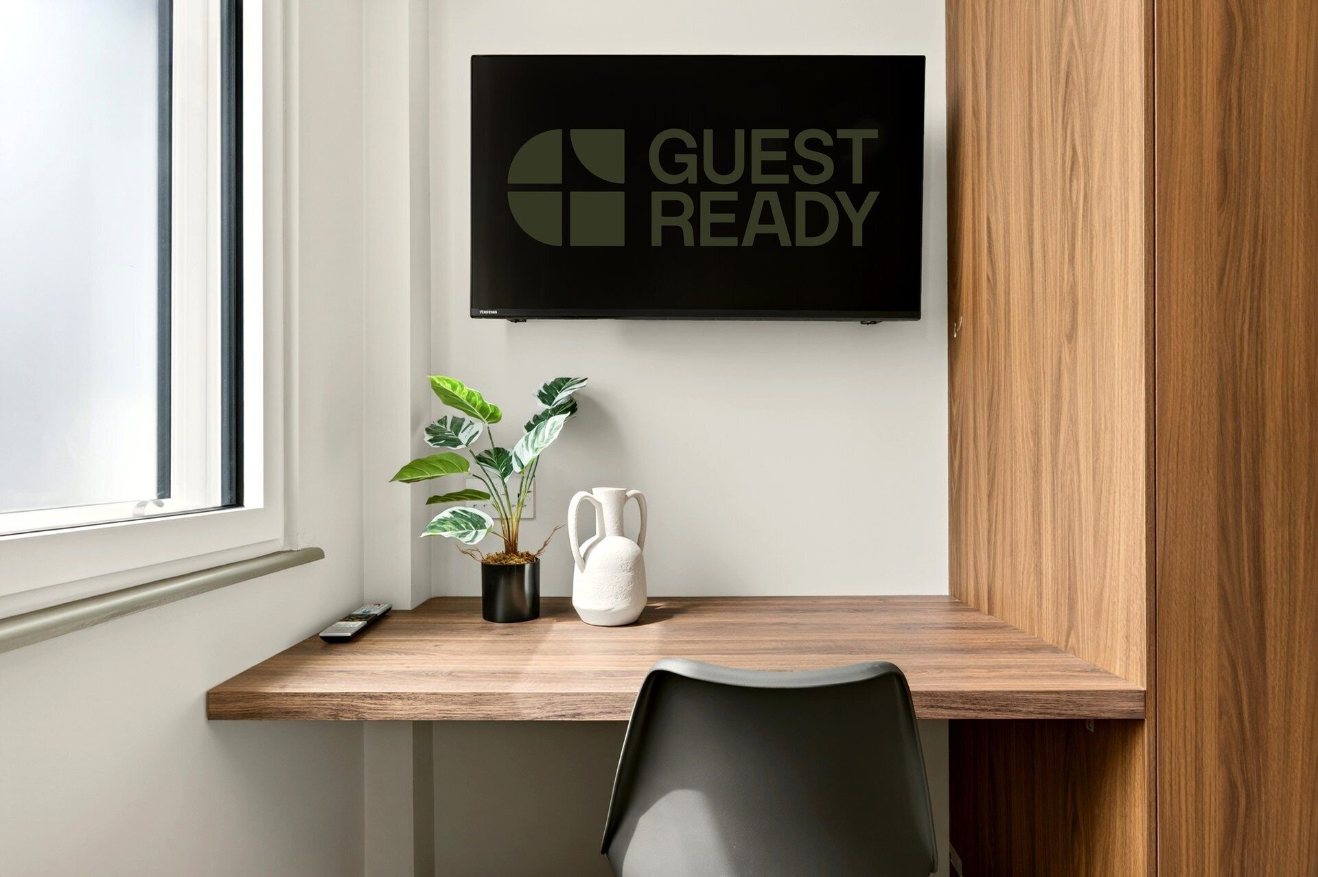 Habitación de hotel con un escritorio de madera, una planta en maceta, un jarrón blanco y un cartel que dice 'GUEST READY'.