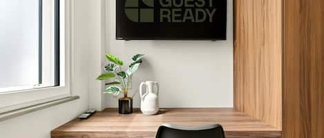 Habitación de hotel con un escritorio de madera, una planta en maceta, un jarrón blanco y un cartel que dice 'GUEST READY'.