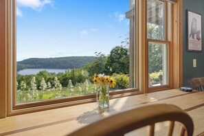 A room with a large window, a wooden table, and a vase with flowers. - Secluded Eastern Maine Mountain House With Breathtaking Views. (Orland)