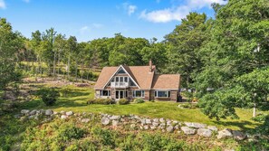 A two-story house with a stone foundation, surrounded by a well-maintained lawn and trees. - Secluded Eastern Maine Mountain House With Breathtaking Views. (Orland)