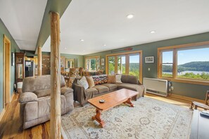 A spacious living room with a large window, a fireplace, and a dining area. - Secluded Eastern Maine Mountain House With Breathtaking Views. (Orland)