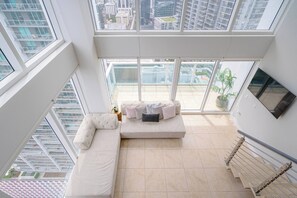 A modern living room with a sectional sofa, a TV mounted on the wall, and a large window offering a city view. - Lovely 2BR Loft in Heart of Brickell w/pool and parking (Miami)