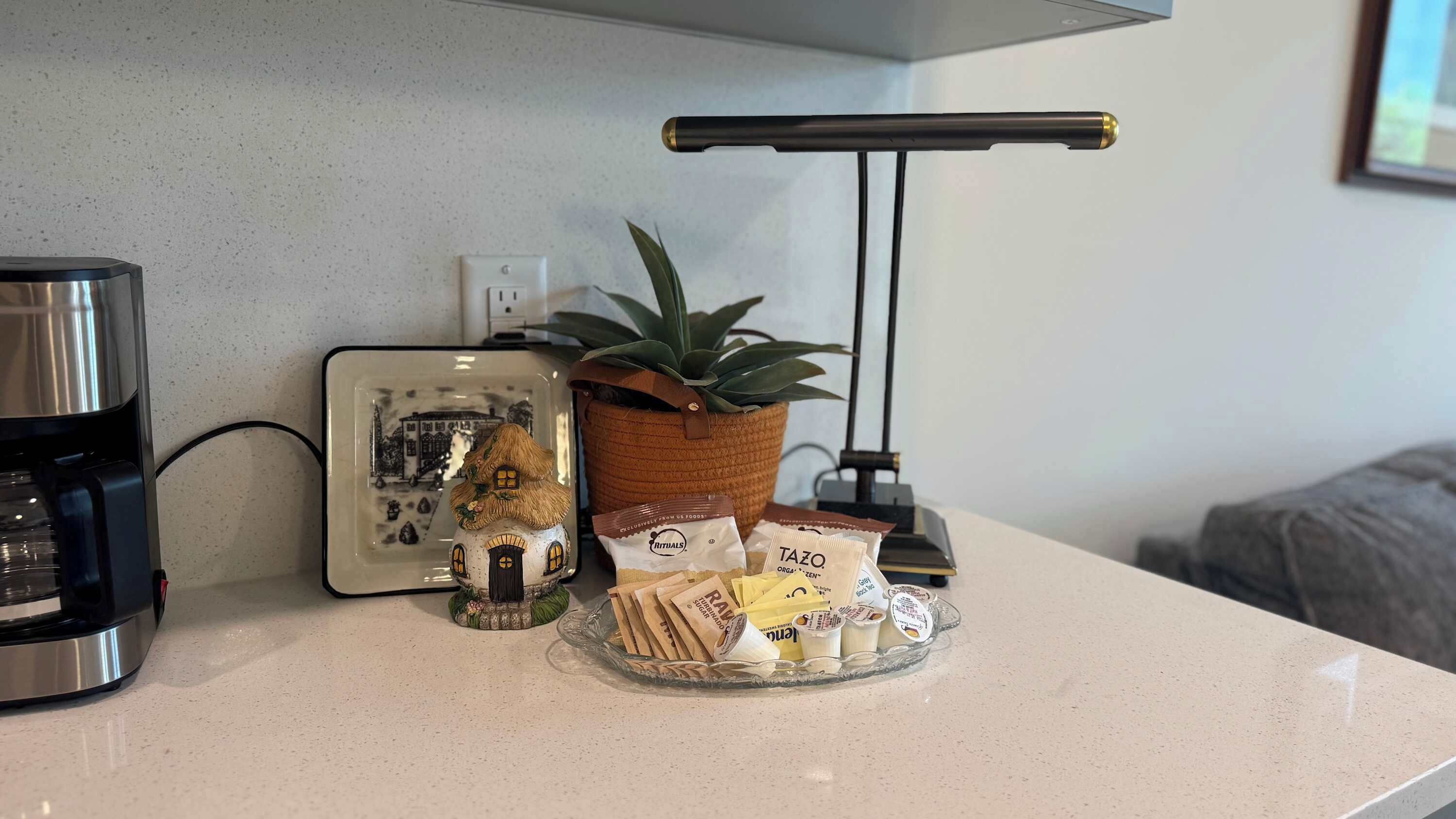 Un comptoir de cuisine avec une machine à café, un tableau encadré, une plante en pot et un plat en verre contenant des aliments.