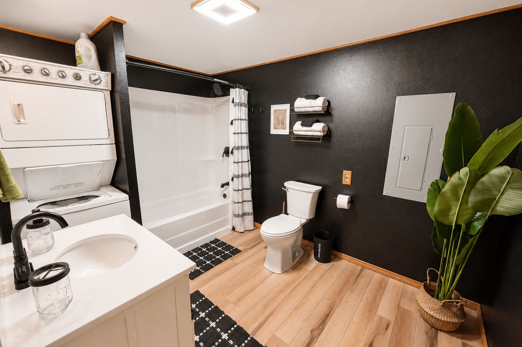 Un baño moderno con lavamanos, inodoro y ducha blancos. Incluye una estufa blanca, una cortina de ducha con estampado blanco y negro, y una planta en maceta.