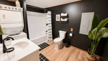 A modern bathroom with a white sink, toilet, and shower. There is a white stove, a black and white patterned shower curtain, and a potted plant.