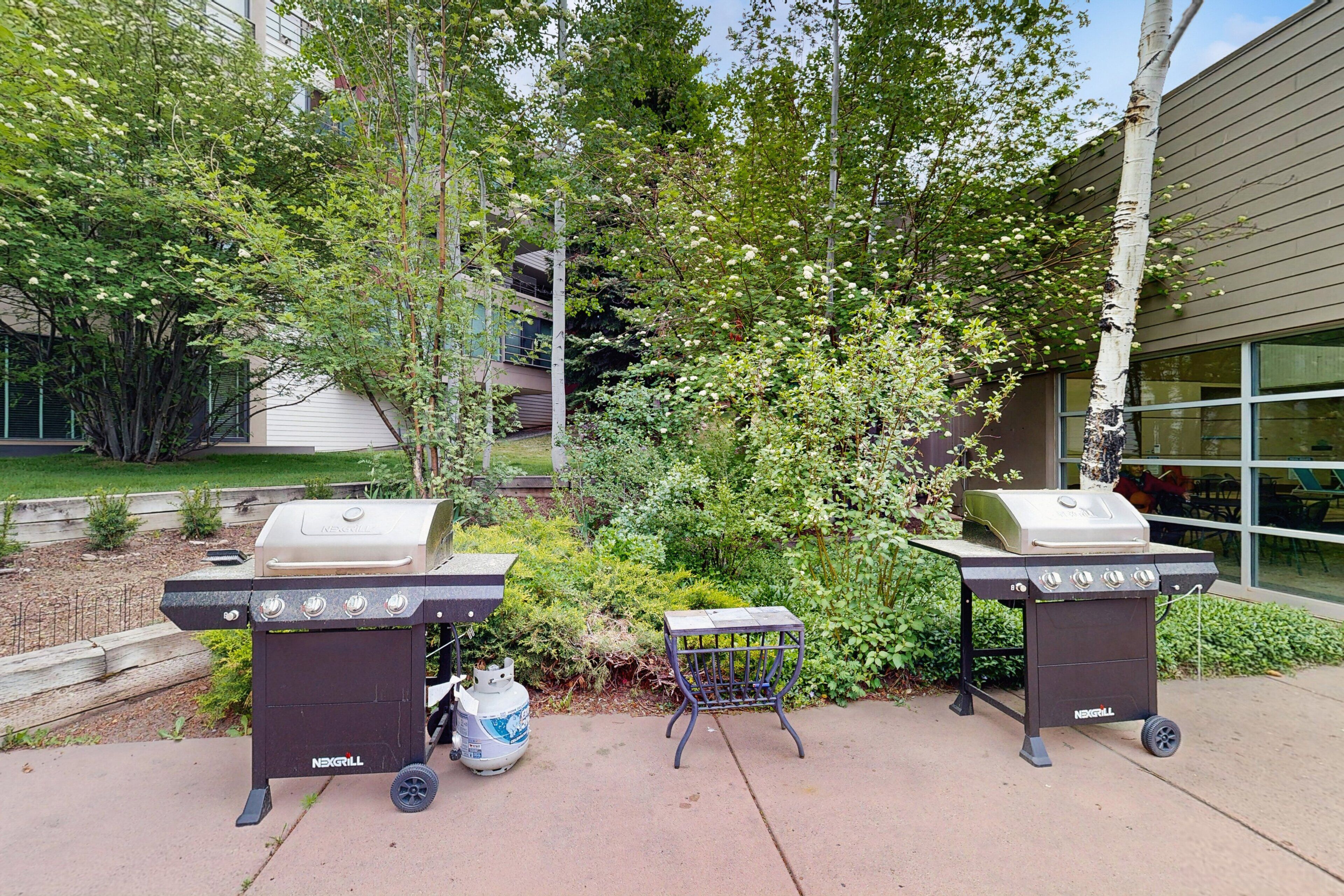 Twee buitenbarbecues, een draagbare tafel en een gasfles in een aangelegde omgeving met bomen en een gebouw op de achtergrond.