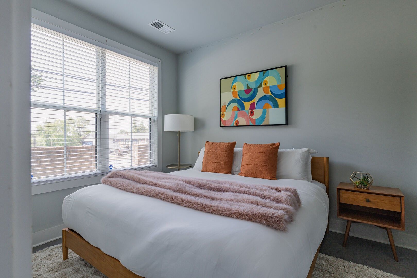 A bedroom with a large bed, a bedside lamp, a nightstand, and a colorful abstract painting on the wall.