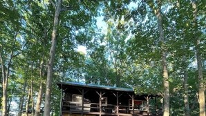 A cabin with a deck, surrounded by trees, and a fire pit in the foreground.