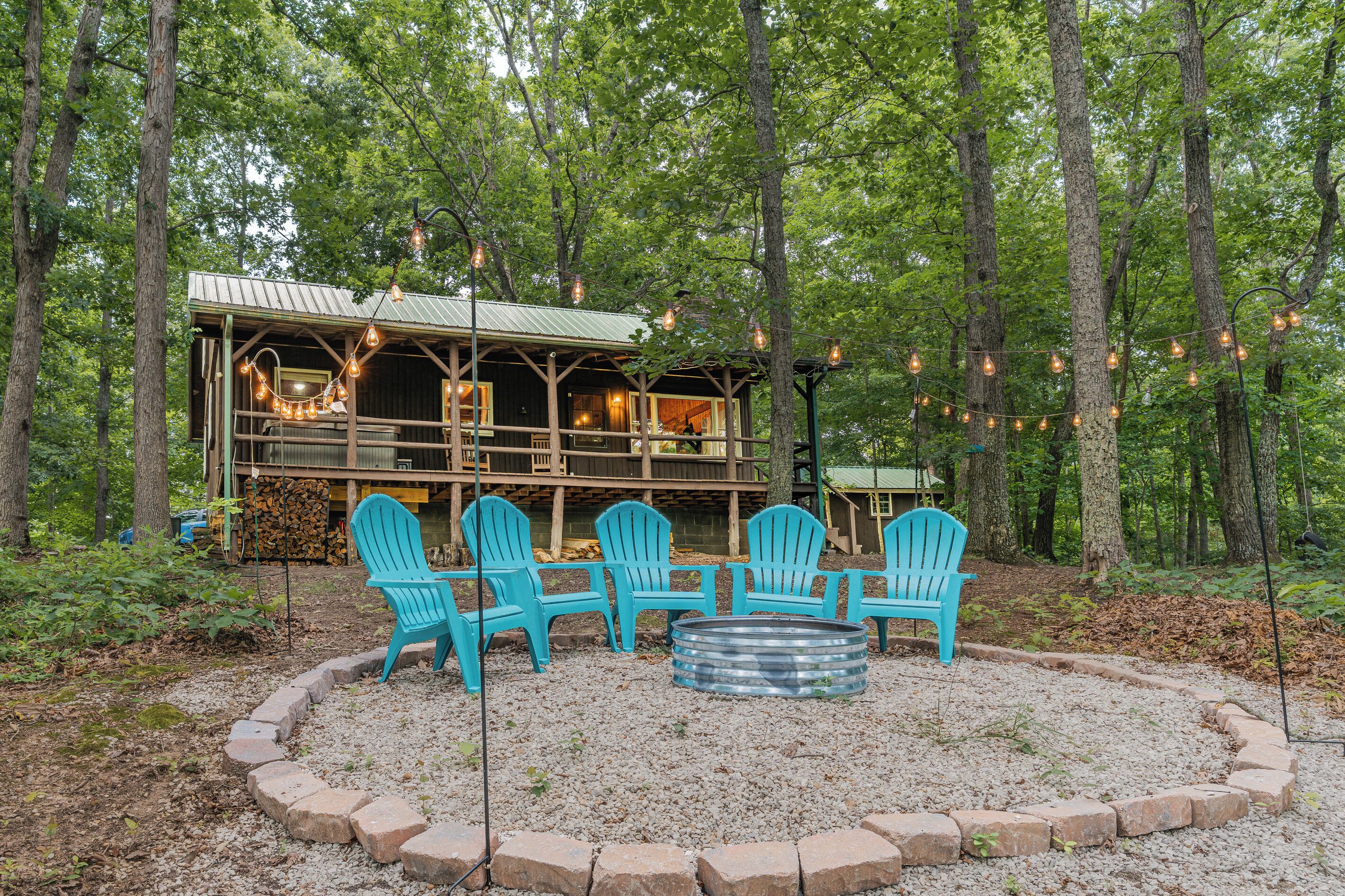 A rustic cabin with a deck, surrounded by trees, and outdoor seating with chairs and a fire pit.