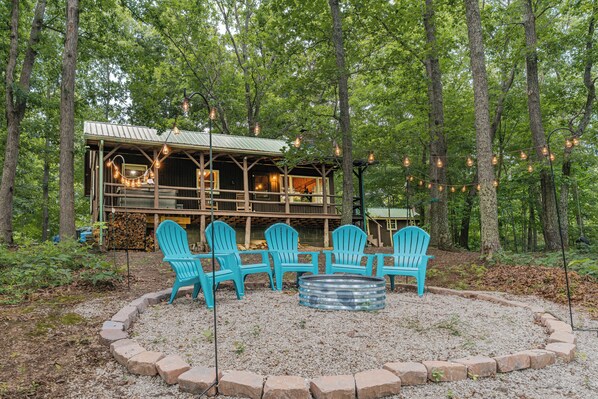 A rustic cabin with a deck, surrounded by trees, and outdoor seating with chairs and a fire pit.