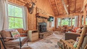 A log cabin interior with a fireplace, wooden furniture, and large windows.