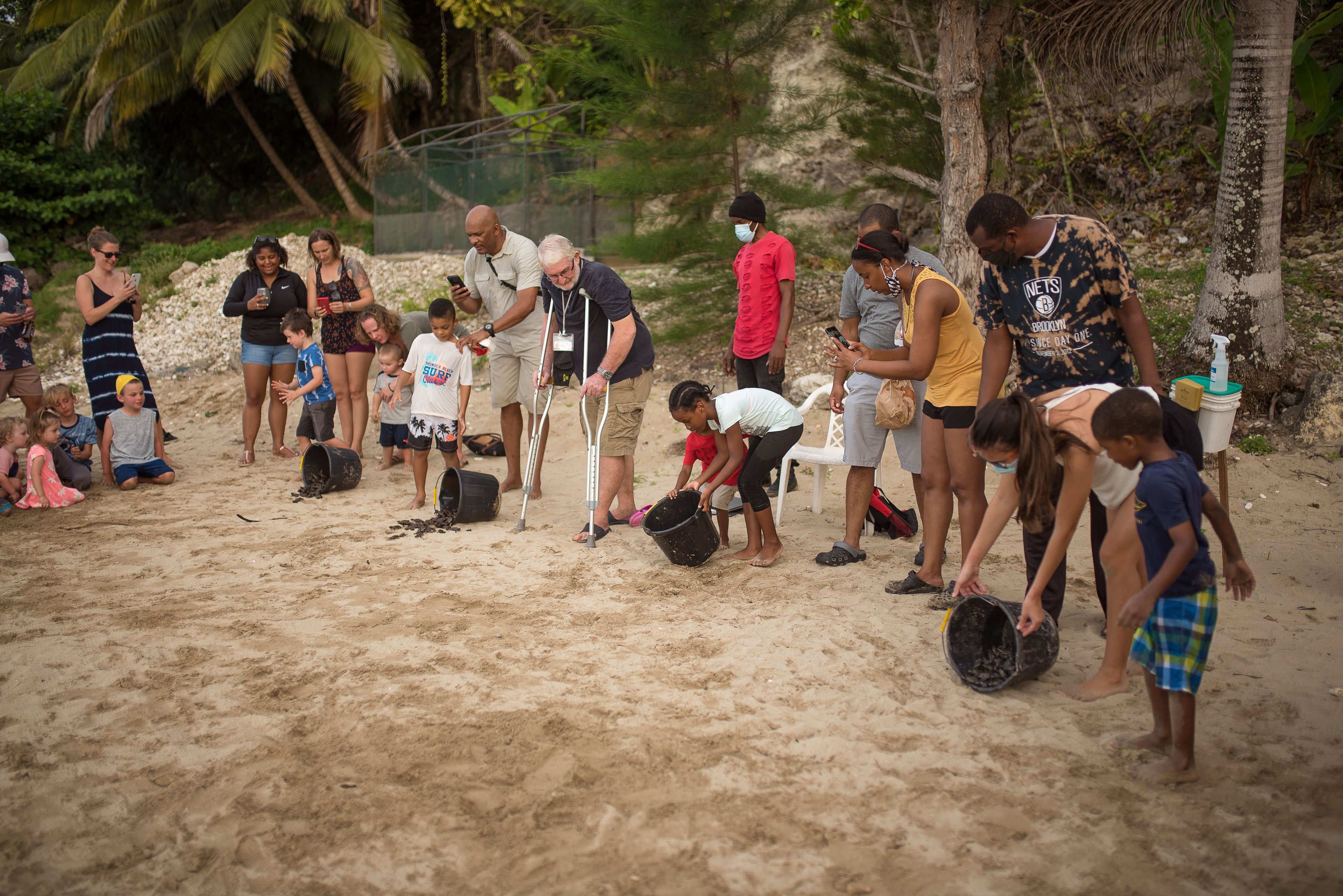 A group of people on a sandy beach with some using crutches, others holding buckets, and a few children playing.