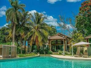 A pool area with lounge chairs, a building with large windows, and palm trees. - Bessa | Ocean View Villa | 2BR, Seaside Pool ➠ U61 (Oracabessa)
