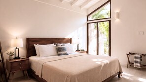 A bedroom with a large bed, two bedside tables, a nightstand, a wooden bench, and a view of greenery through a large window. - Bessa | Ocean View Villa | 2BR, Seaside Pool ➠ U61 (Oracabessa)