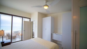 Quarto de hotel com uma cama, um ventilador de teto, banheiro com pia e vista para o mar através de uma porta de correr.