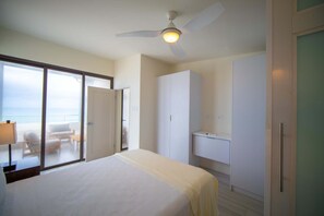 Ein Hotelzimmer mit einem Bett, einem Deckenventilator, einem Badezimmer mit Waschbecken und Blick auf den Ozean durch eine Schiebetür.