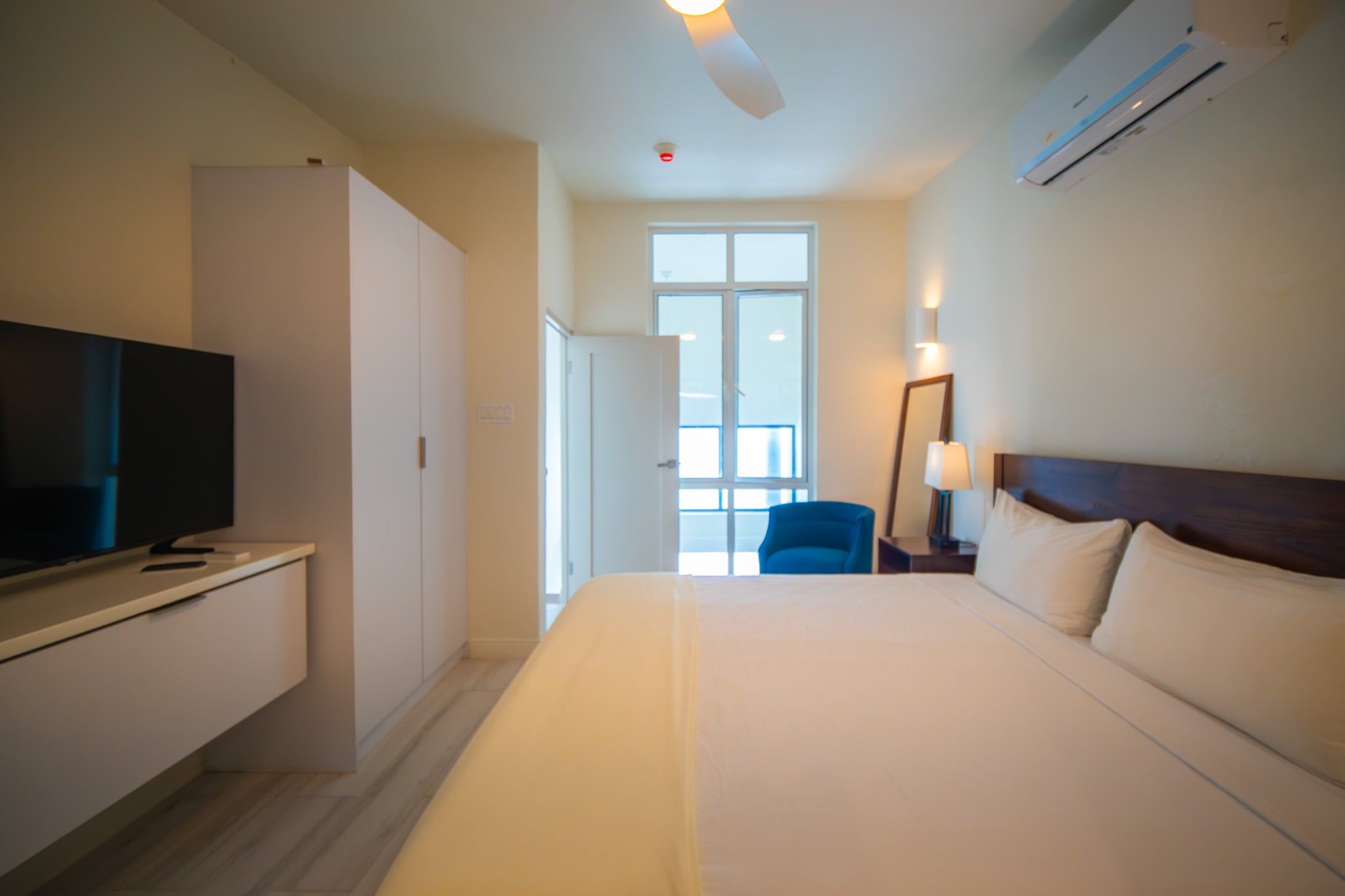 Representative Bessa room detail; layout, furnishings and bedding may vary by unit. A blue chair sits near the bedroom windows, alongside the bed, wardrobe and wall-mounted TV.