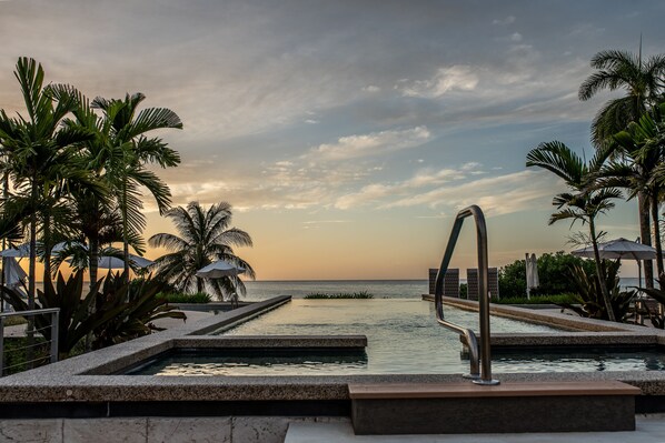 A pool area with a view of the sea, surrounded by palm trees and umbrellas.