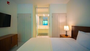 A hotel room with a bed, a TV, a bedside table with a lamp, a wardrobe, and a bathroom visible through an open door. - Bessa | Beachfront Studio | Seaside Pool ➠ U77 (Oracabessa)