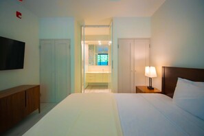 Quarto de hotel com cama, televisão, mesa de cabeceira com abajur, guarda-roupa e banheiro visível através de uma porta aberta.