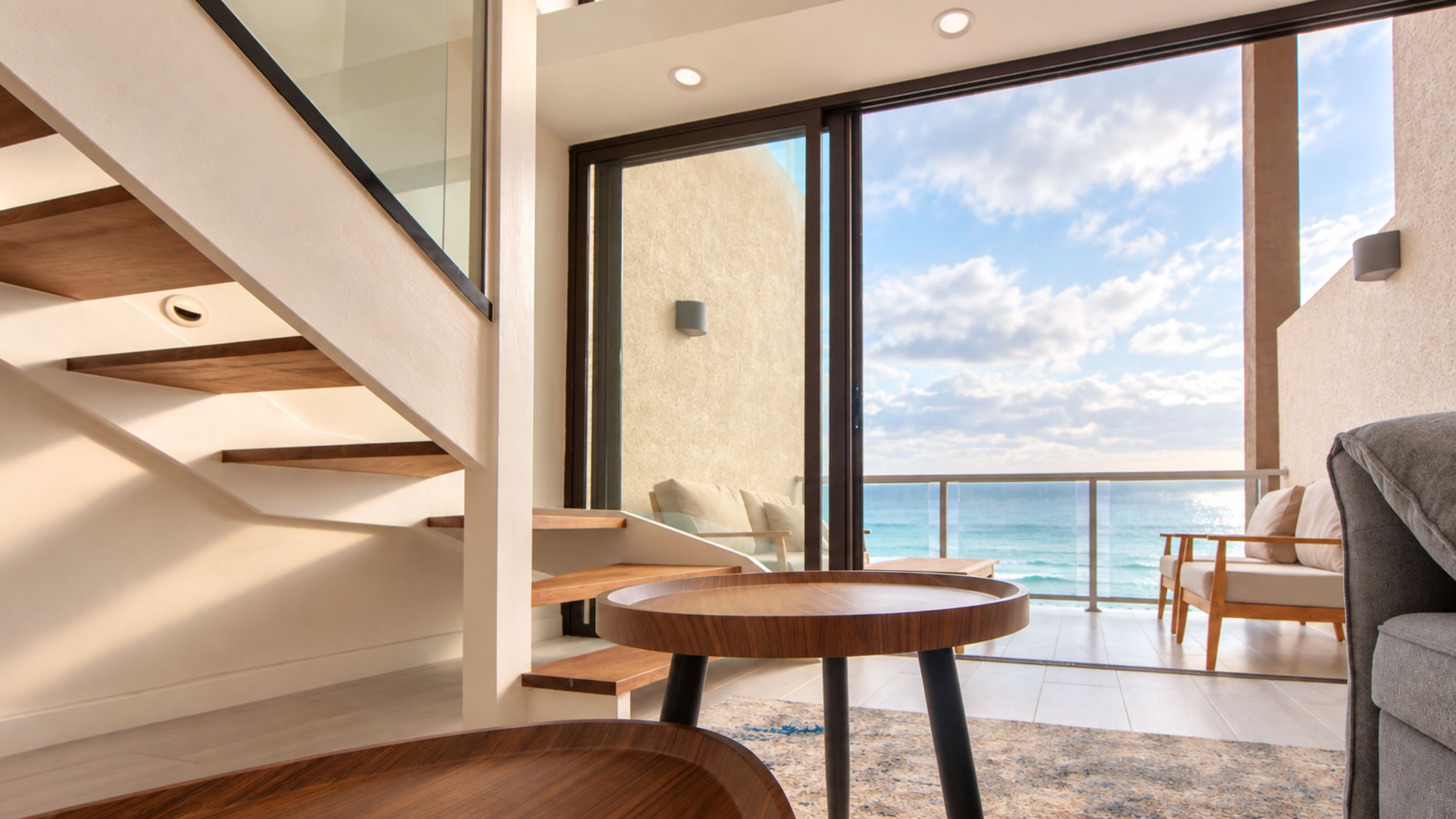 A modern interior with a staircase, a balcony with a view of the sea, and a seating area with a table.