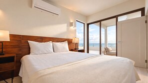 Une chambre d’hôtel avec un grand lit, une tête de lit en bois, une table de chevet, une lampe et une vue sur la mer, visible à travers une porte coulissante.