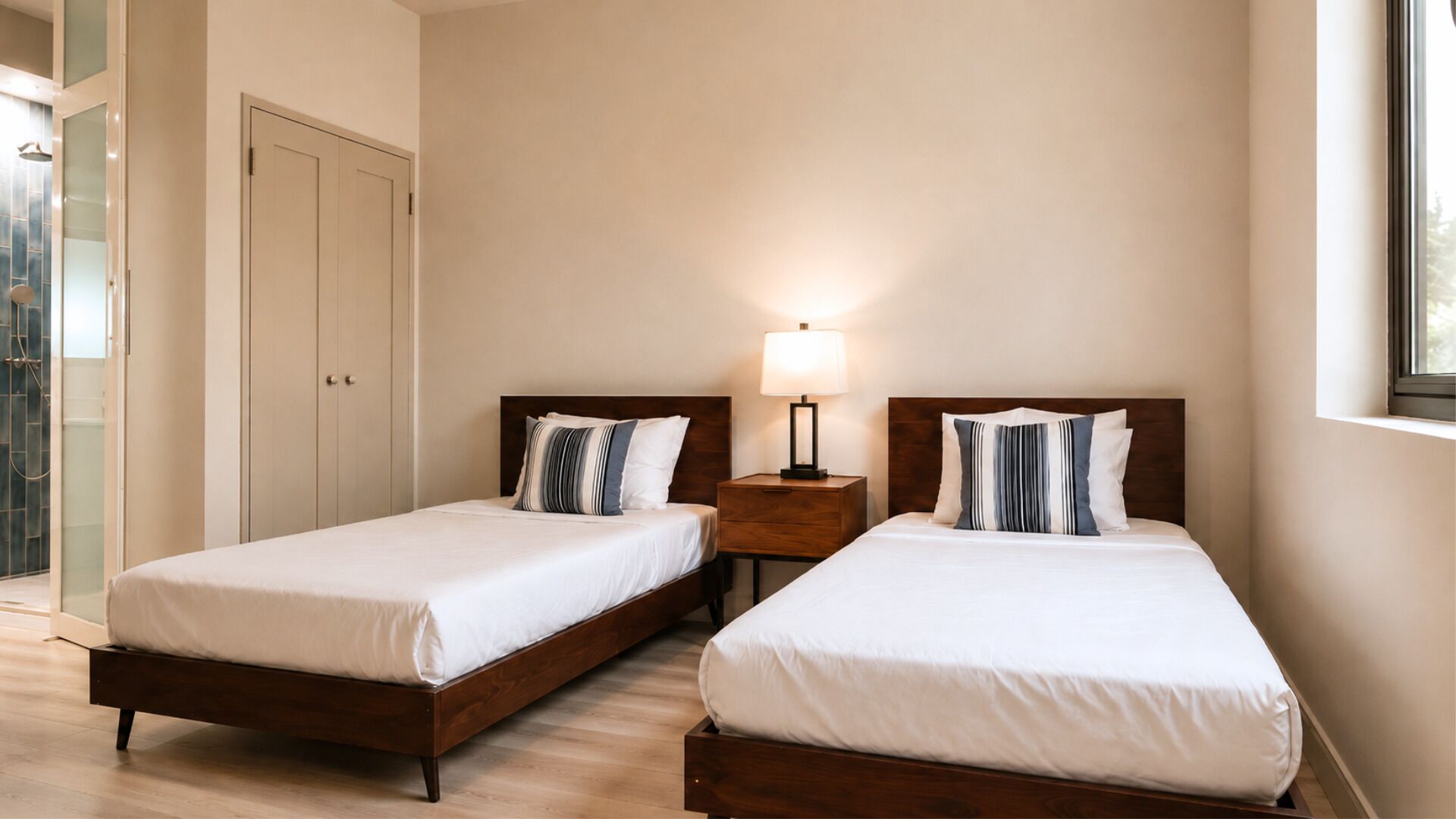 Ein Hotelzimmer mit zwei Einzelbetten, einem Nachttisch mit Lampe, einem Kleiderschrank und einem Fenster.