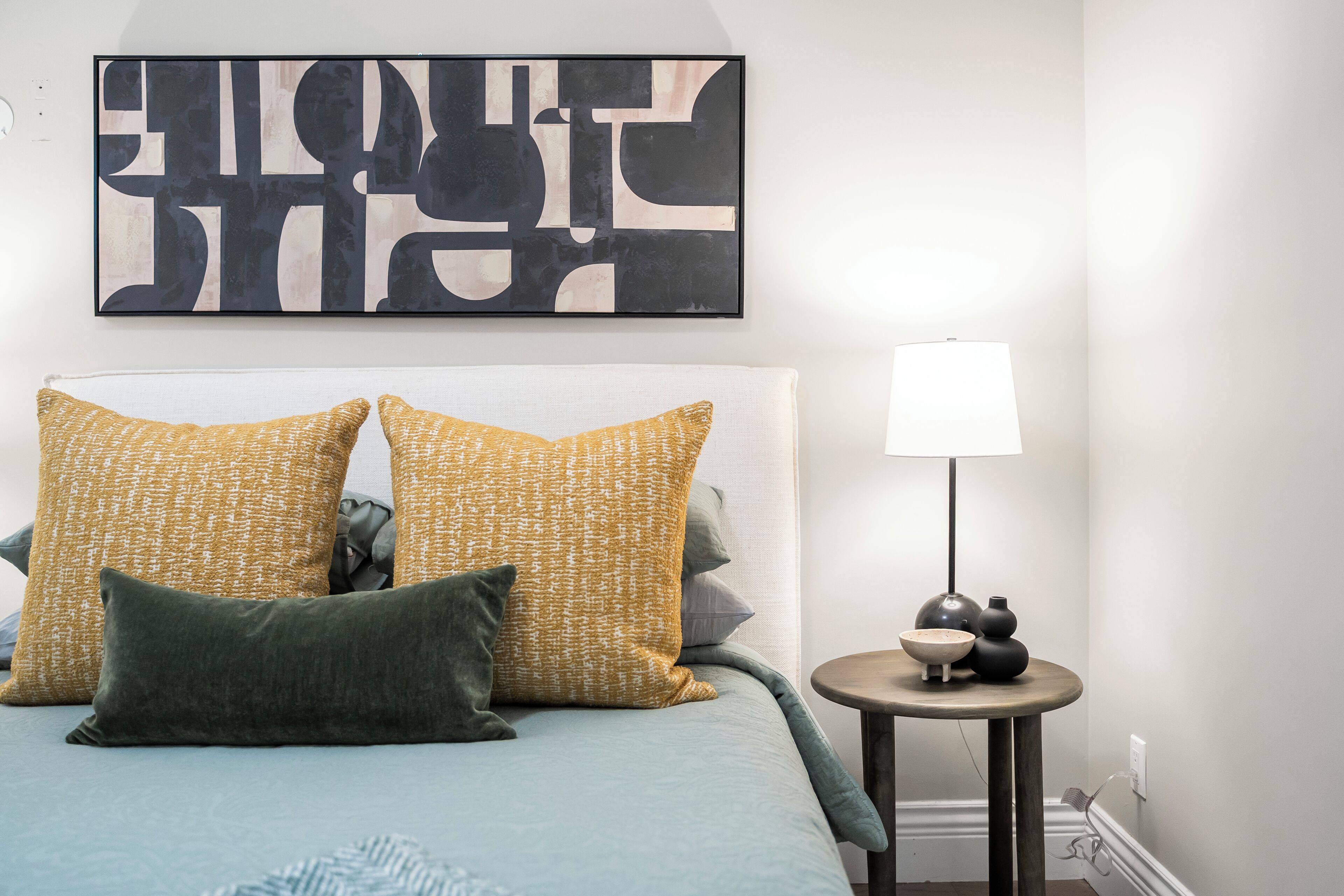 A bedroom with a bed, pillows, a bedside table with a lamp, and a wall art piece.