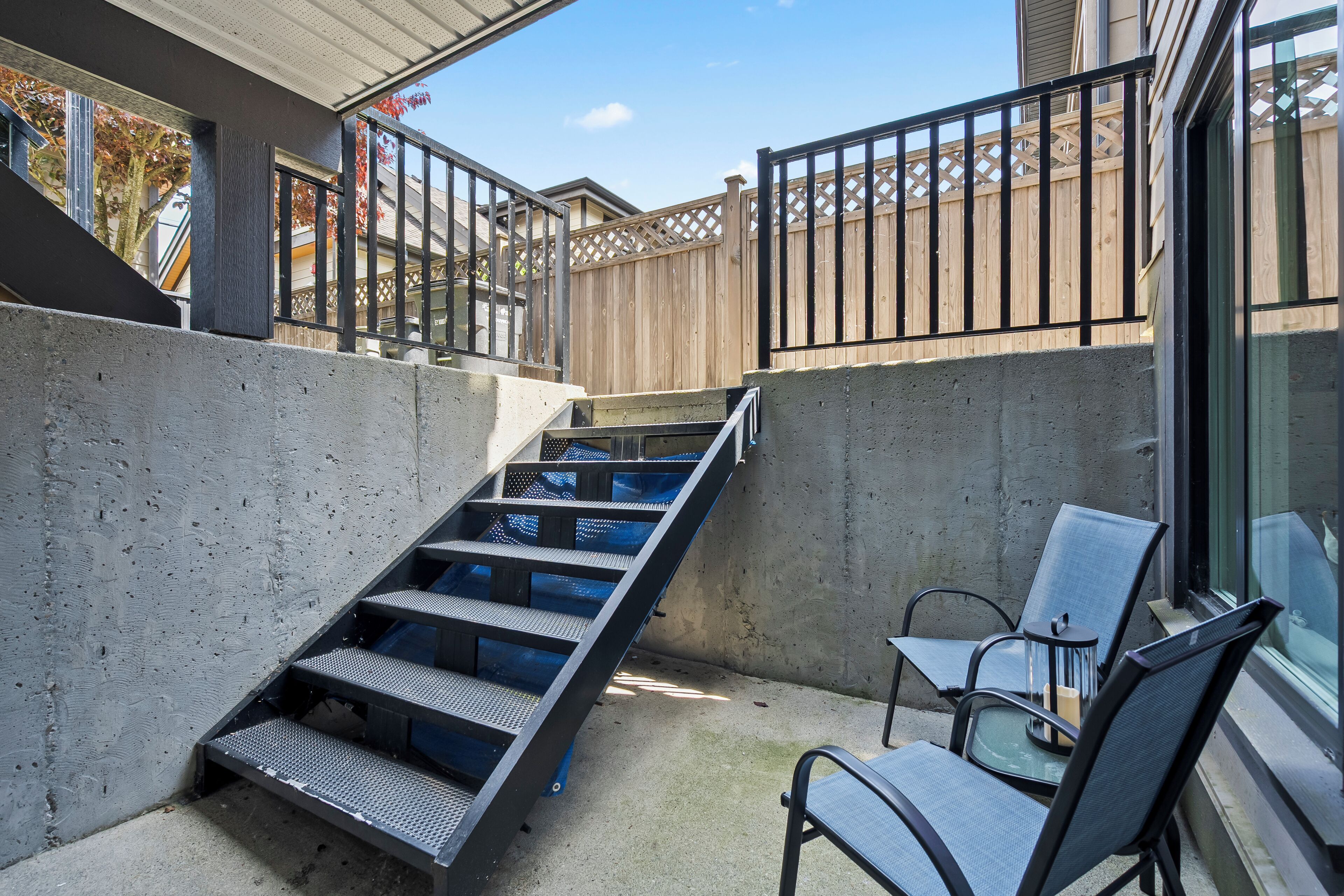 Una escalera de concreto que lleva a un balcón, una mesa de vidrio con sillas y una valla de madera.