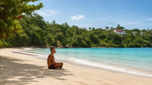 A beach with a person sitting on the sand, trees, and a building in the background.