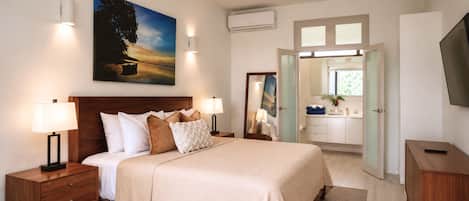 Une chambre d’hôtel avec un grand lit, des lampes de chevet, un tableau, un appareil de climatisation, une salle de bain avec un lavabo et un miroir, et un téléviseur.