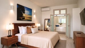 Ein Hotelzimmer mit einem großen Bett, Nachttischlampen, einem Gemälde, einer Klimaanlage, einem Badezimmer mit Waschbecken und Spiegel sowie einem Fernseher.