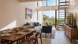 A spacious living area with a dining table set for four, a comfortable sofa, and a view of the outdoors through large windows. - Bessa | Riverfront 2BR Loft, Private Roof Terrace ➠ U11 (Oracabessa)
