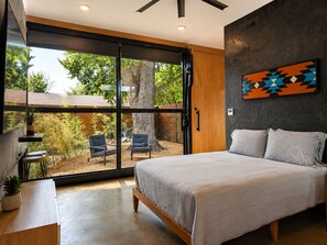 A bedroom with a large sliding glass door leading to a patio, a bed with a headboard, a wall-mounted abstract painting, and a small table with a plant. - LA ROCA - Boutique Fayetteville Studio (Fayetteville)