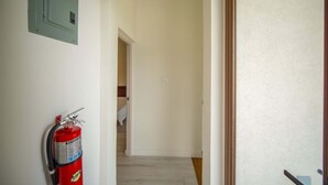 A hallway with a fire extinguisher mounted on the wall, a door at the end, and a light switch on the wall. - Bessa | Riverfront 2BR | River, Sea & Pool ➠ U4 (Oracabessa)