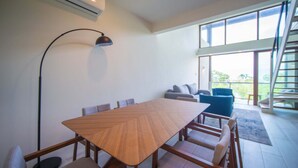 A modern dining area with a large wooden table, cushioned chairs, a floor lamp, and a staircase leading to an outdoor balcony. - Bessa | Riverfront 2BR | River, Sea & Pool ➠ U4 (Oracabessa)