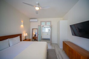 Ein Hotelzimmer mit Bett, Deckenventilator, Fernseher und einem Badezimmer mit Spiegel, sowie einer Tür zu einem weiteren Raum.