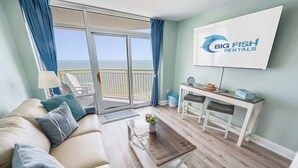 Una sala de estar moderna con un sofá, una mesa de centro y un escritorio con dos sillas. Un ventanal ofrece vistas al mar.