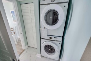 A laundry room with a Samsung washer and dryer, a white door, and a view of a room with a window. - Bay Watch 1518 Direct Oceanfront, Tiki Bar & Pools (North Myrtle Beach)