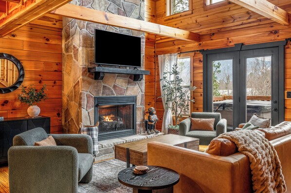 A cozy log cabin interior with a stone fireplace, comfortable seating, and a large window. - Highland Cabin w/ Mountain Views in Chalet Village (Gatlinburg)