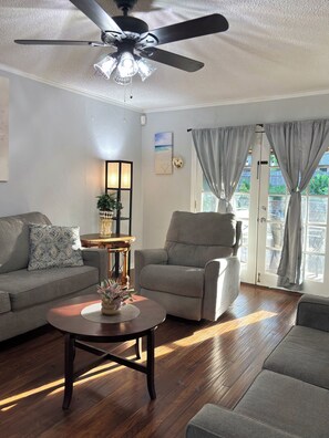 Ein Wohnzimmer mit Deckenventilator, einer Couch, einem Sessel, einem Couchtisch und Blick durch eine Tür.