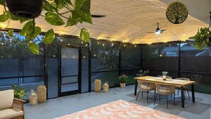 Un patio techado al aire libre con una mesa y sillas, luces colgantes y plantas en macetas.