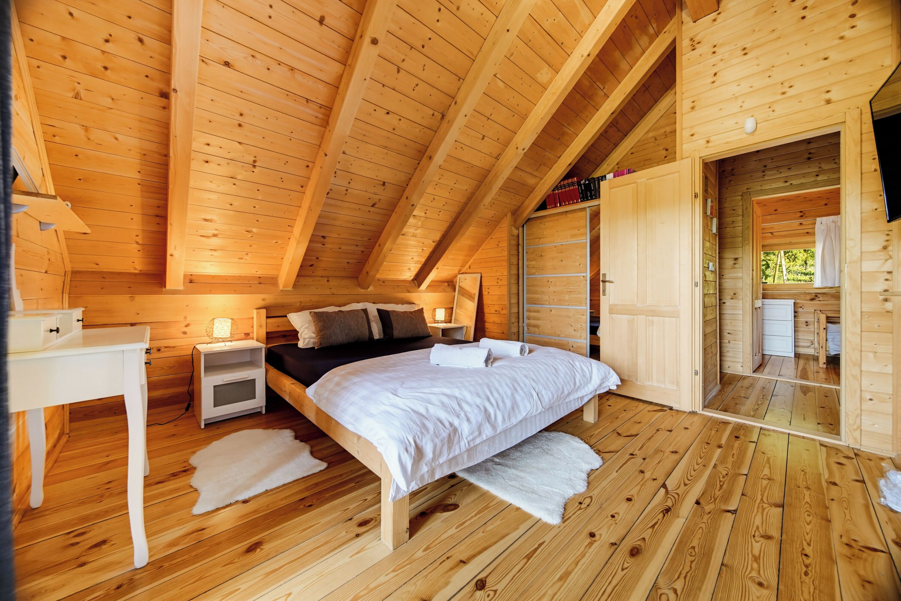 Un dormitorio de cabaña de madera con cama, mesitas de noche, un escritorio y un baño visible a través de una puerta abierta.