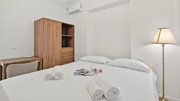 Ein Hotelzimmer mit einem Bett, einem Holzschrank, einem Schreibtisch, einer Lampe und einer Zeitschrift auf dem Bett.
