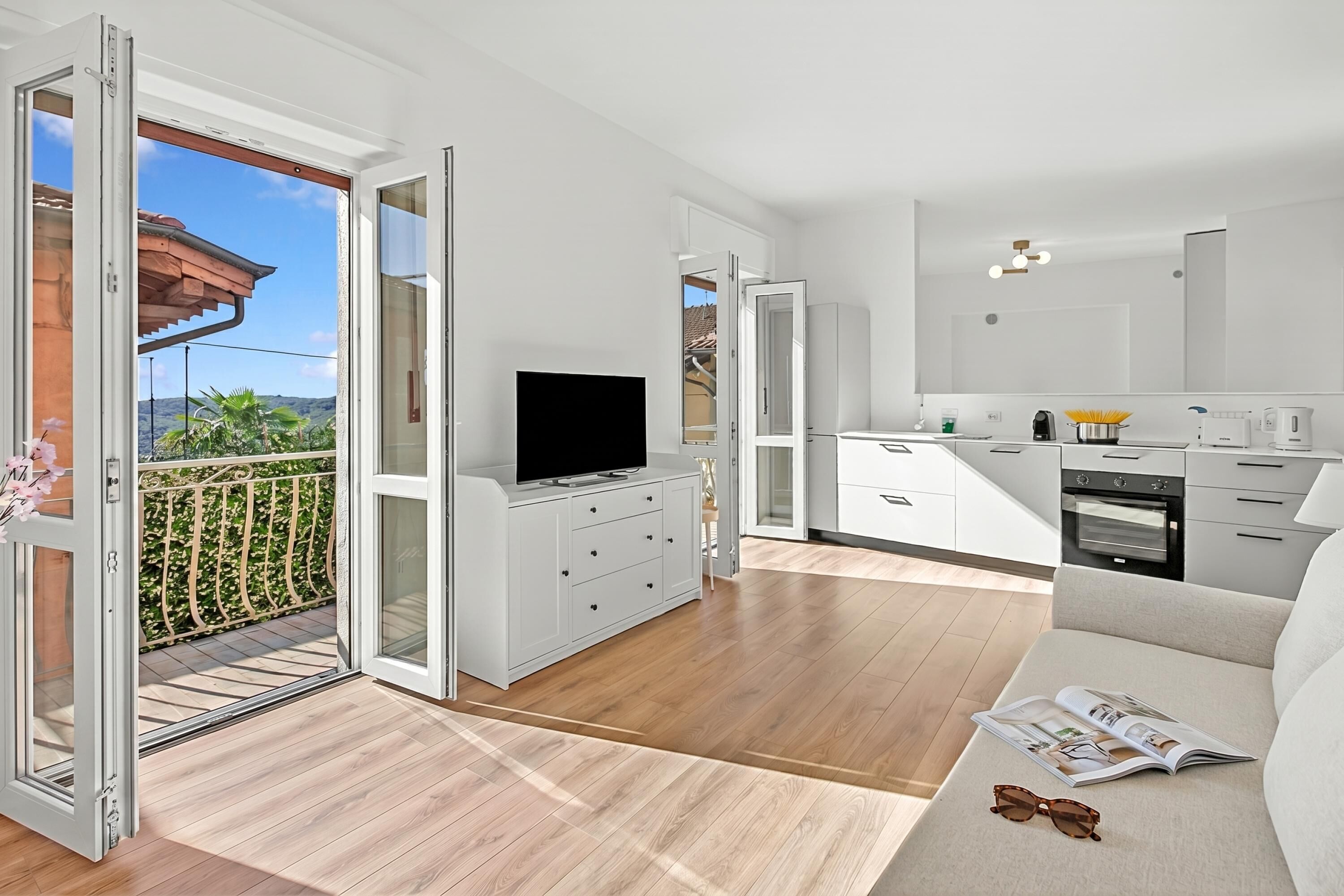 A modern apartment with a balcony, a flat-screen TV, a kitchen with appliances, and a sofa with a magazine.