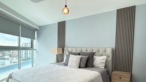 Une chambre moderne avec un grand lit, une table de chevet, une lampe et une vue sur la ville grâce à de grandes fenêtres.