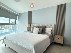 Ein modernes Schlafzimmer mit einem großen Bett, einem Nachttisch, einer Lampe und Blick auf die Stadt durch große Fenster.