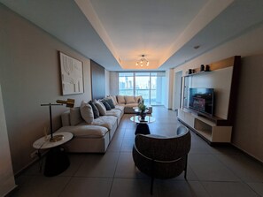 Ein modernes Wohnzimmer mit einer Eckcouch, einem runden Couchtisch, einem Fernseher und einem großen Fenster mit Blick auf die Stadt.