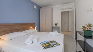 Quarto de hotel com uma cama, mesa de cabeceira com uma revista, planta em vaso e guarda-roupa.